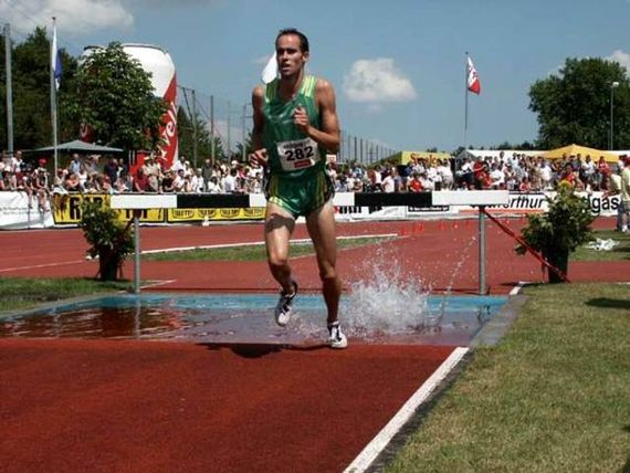 Ein Wassergraben auf der roten Tartarbahn bei einem Wettkampf. Ein Sportler springt über diesen Graben, der mit Wasser gefüllt ist.