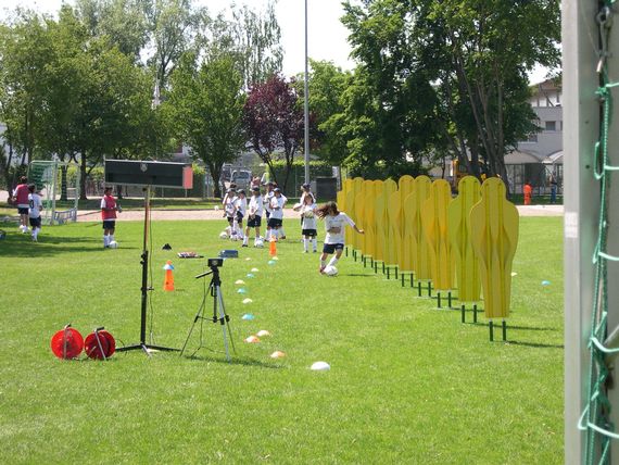 Auf der Wiese sind gelbe Figuren aufgestellt. Die kleine Kinder müssen versuchen mit dem Ball am Fuss um diese Figuren zu spielen.