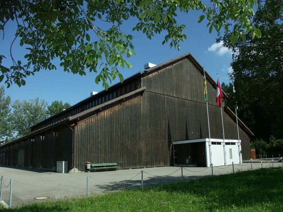 Die Festhalle Rüegerholz fotografiert unter einem Baum hervor, mit Blätter am oberen Bildrand. 