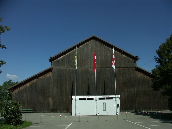 Die Front der Festhalle Rügerholz mit dem Eingangsbereich. Davor stehen drei Fahnenmasten mit der Frauenfelder-, Thrugauer- und Schweizerfahne.