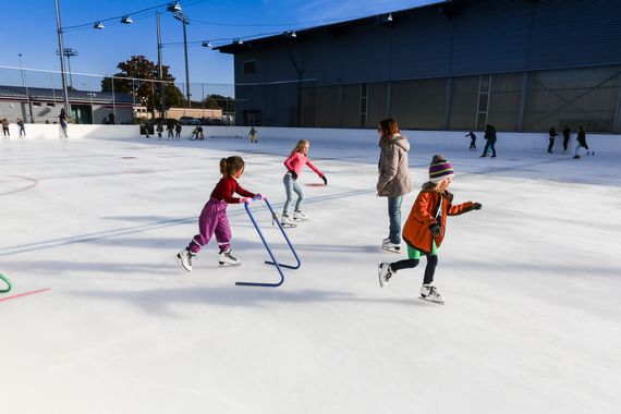 Kleine Kinder auf dem Eis. Eines davon ist mit einer Schlittschuhhilfe unterwegs, welche vorne gehalten werden kann.
