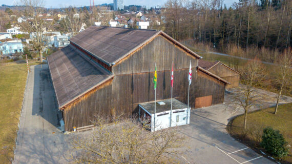 Eine Drohnenaufnahme der Festhalle. Im Vordergrund ist der Eingangsbereich und die Fahnenmasten zu sehen. 